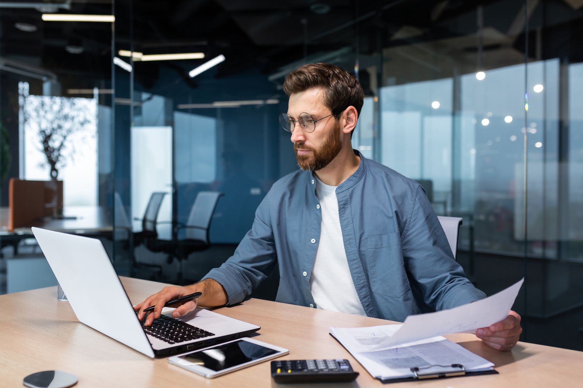 Man with beard and glasses working on laptop and papers in modern office.