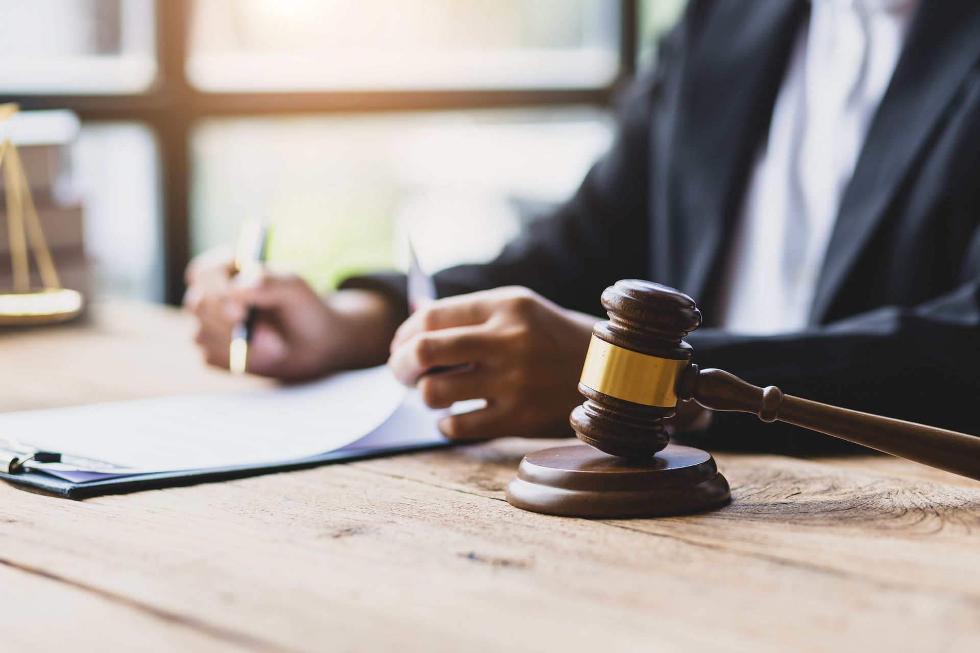 A person in a suit writing at a desk with a gavel, gold scales, and papers; natural light.