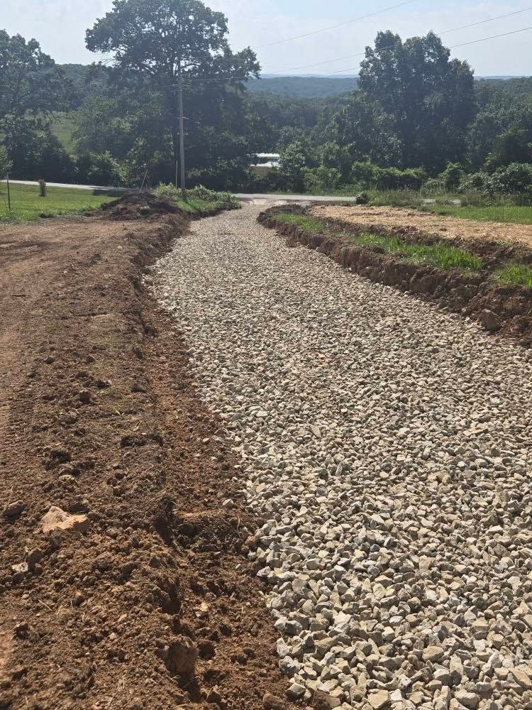 A gravel driveway under construction next to a dirt patch, leading into a wooded area on a sunny day.