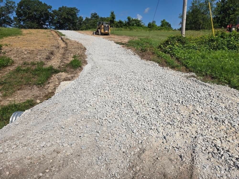 Gravel driveway under construction, small bulldozer in background, trees and grass surround.