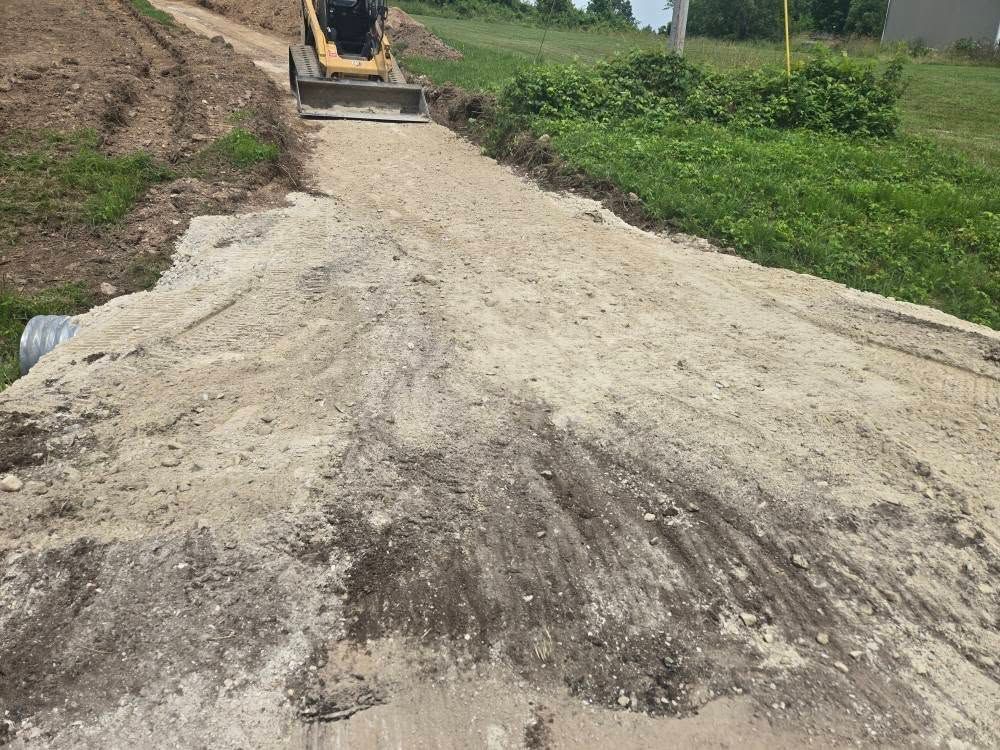 A small bulldozer spreads gravel on a dirt road. Green grass borders the road.