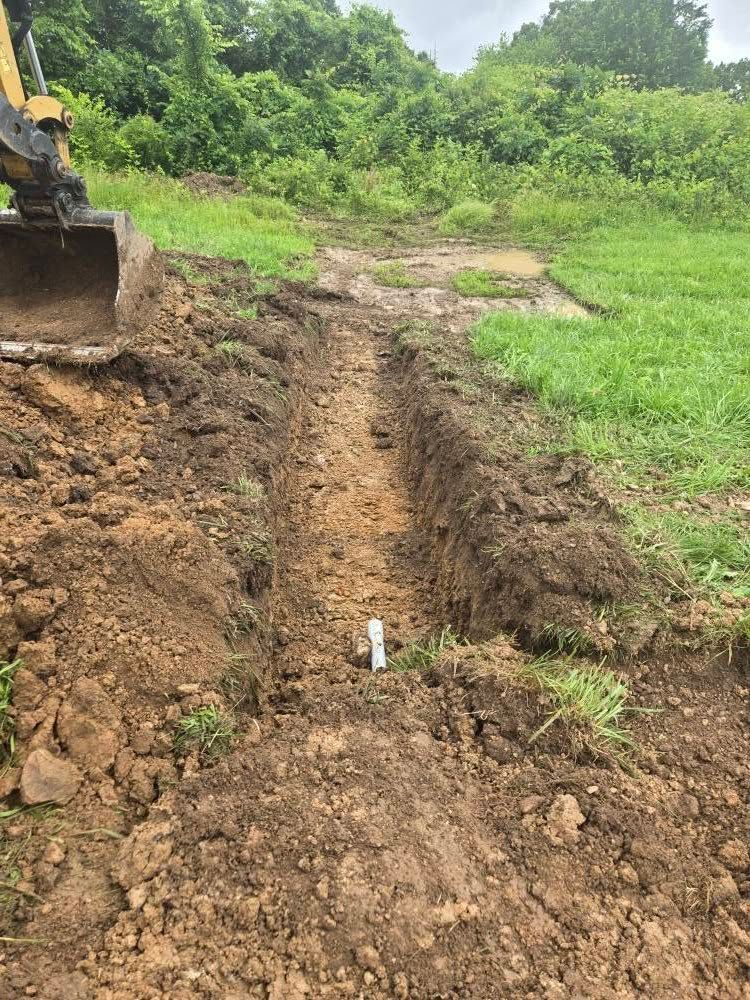 An excavator digging a trench in dirt, revealing a white pipe. Green grass and foliage in background.