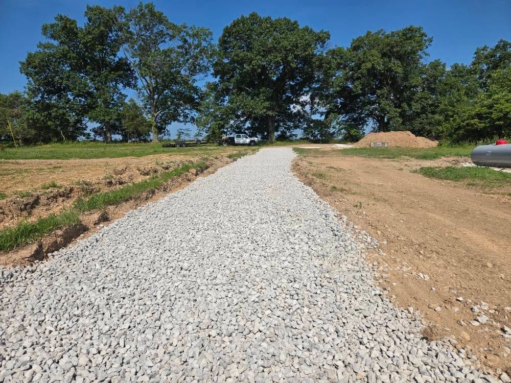 Gravel driveway leading through a rural landscape, lined with dirt and grass. Trees and blue sky in background.