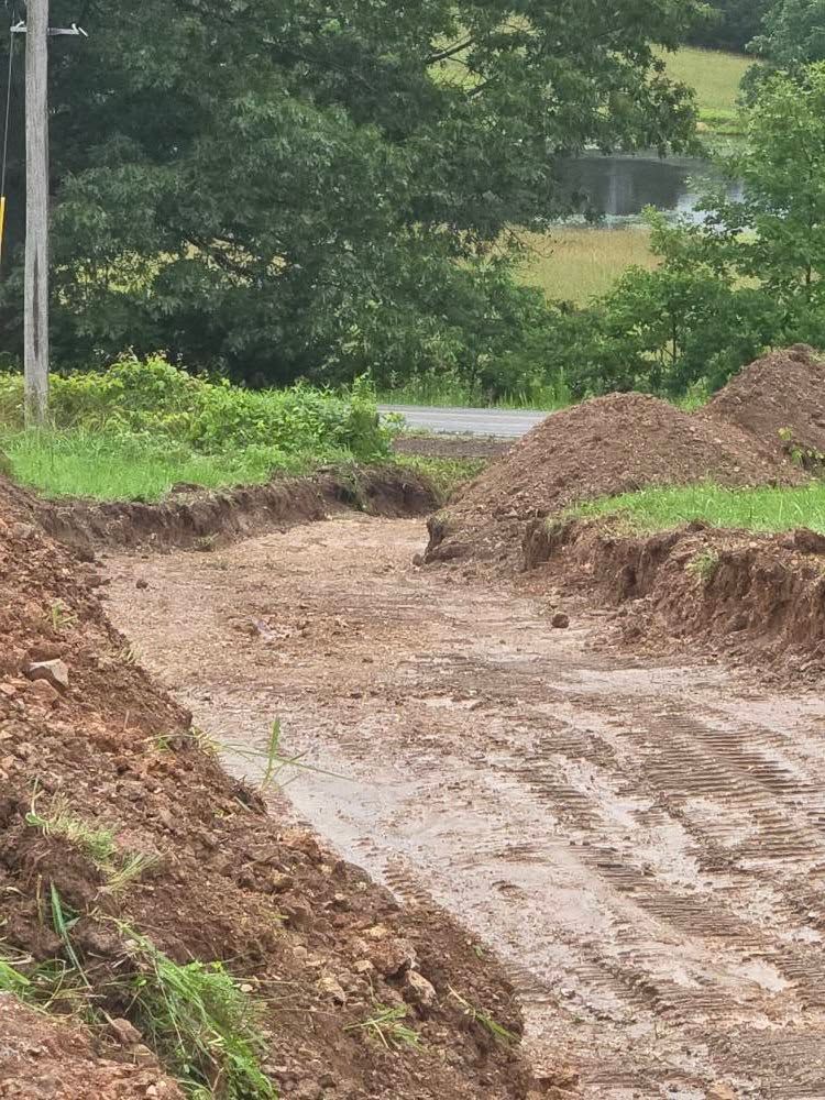 Dirt road under construction, surrounded by piles of dirt, grassy bank, and trees.