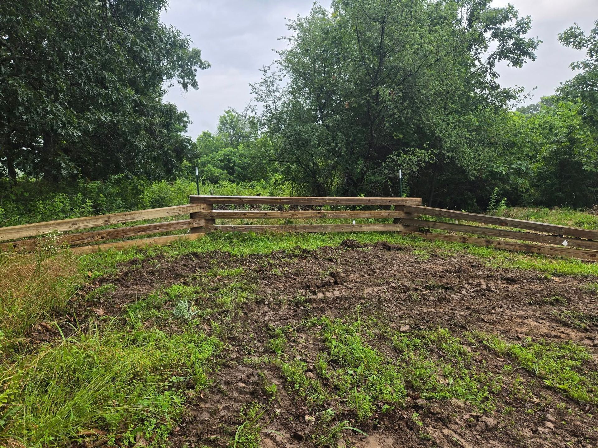 A wooden fence sits atop a muddy hillside, surrounded by green grass and trees, under a cloudy sky.