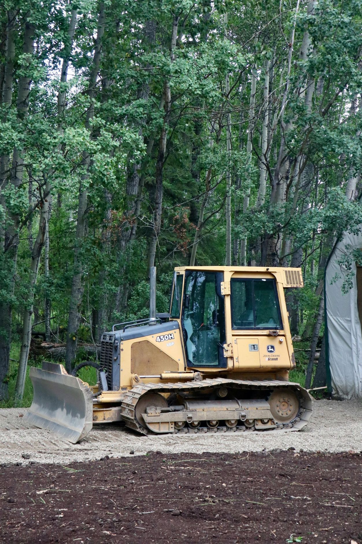 Yellow bulldozer on a gravel surface, set against a backdrop of trees and a white structure.