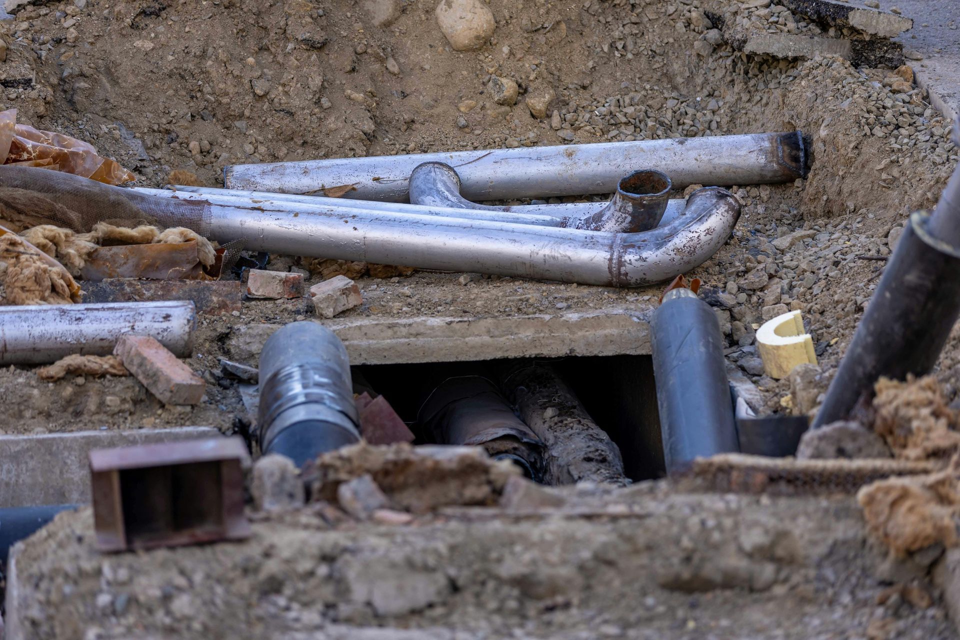 Pipes in an excavated area, some metallic, some plastic, surrounded by dirt and debris.