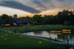 Sunset over a Japanese garden with a pond, boats, pathways, and lit lanterns.