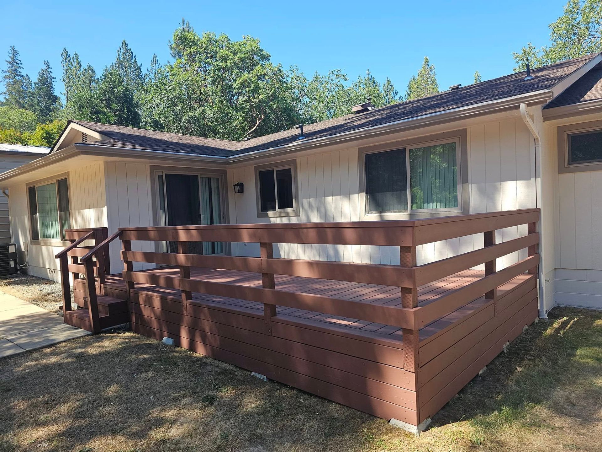 Brown deck attached to a light-colored house with a sliding glass door and windows. Blue sky.
