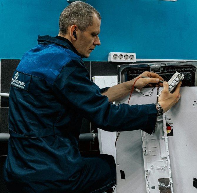 A person wearing a blue work uniform repairs an electronic appliance at a workbench against a blue wall.