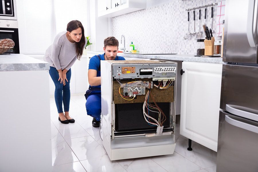 A technician in work coveralls repairs a dishwasher pulled out from under a white kitchen counter, as a person watches.