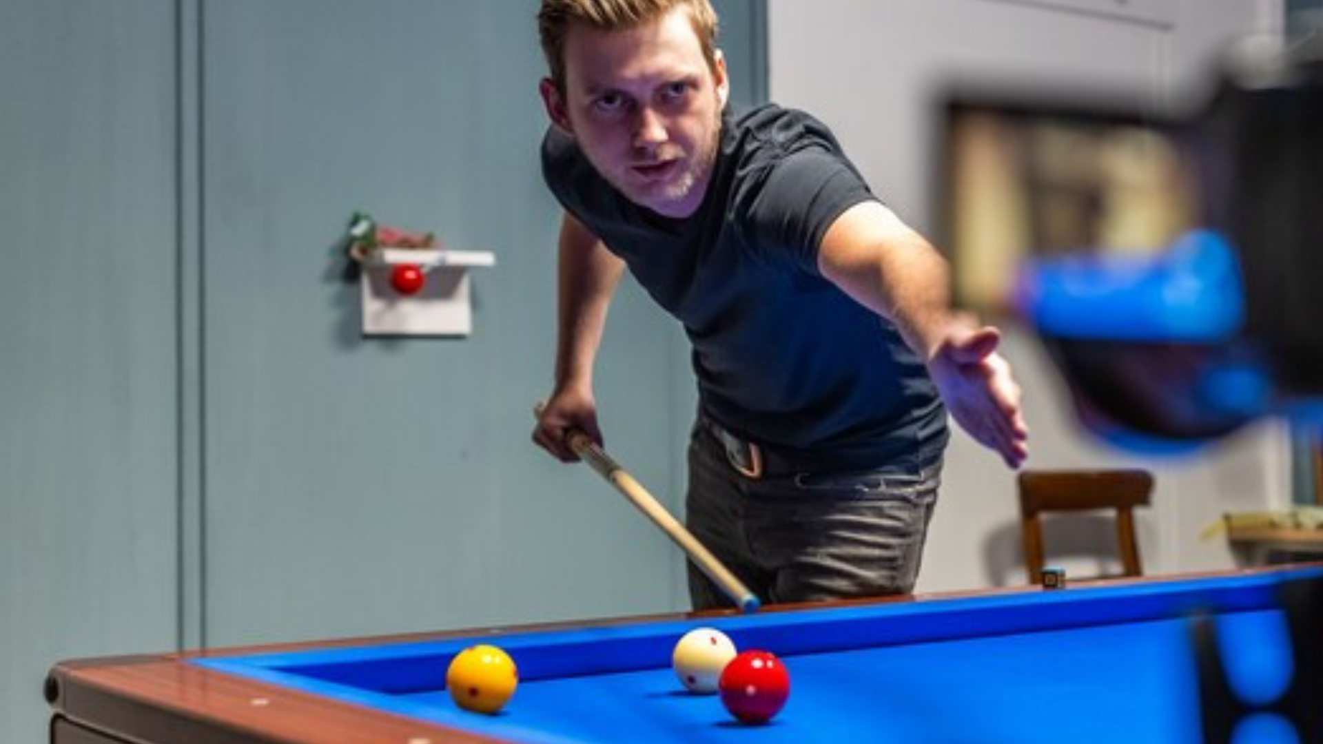 Een man in een zwart shirt richt met een biljartkeu op biljartballen op een blauwe tafel.