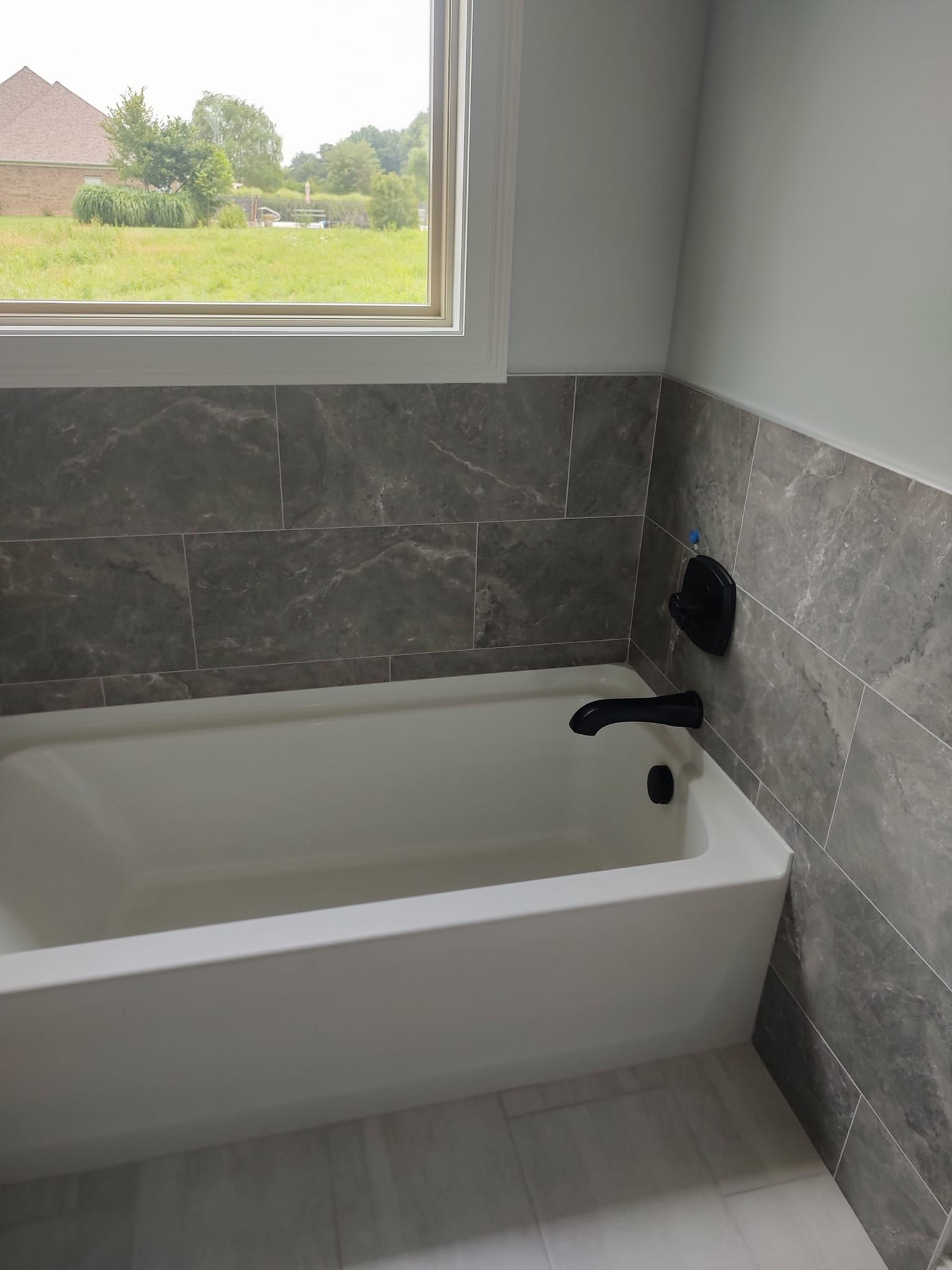 White bathtub with black faucet, against gray marble tile in a bathroom with a window.