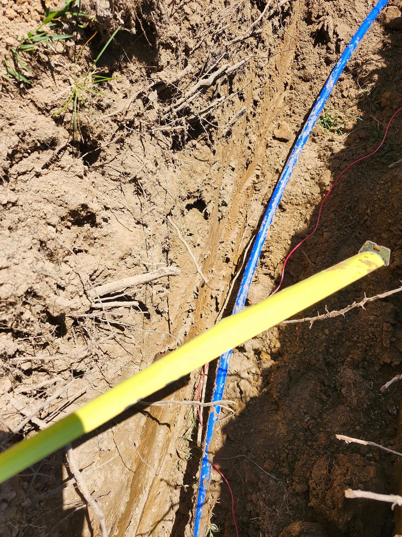 Trench in dirt with blue cable and yellow tape measure, likely for electrical work outdoors.