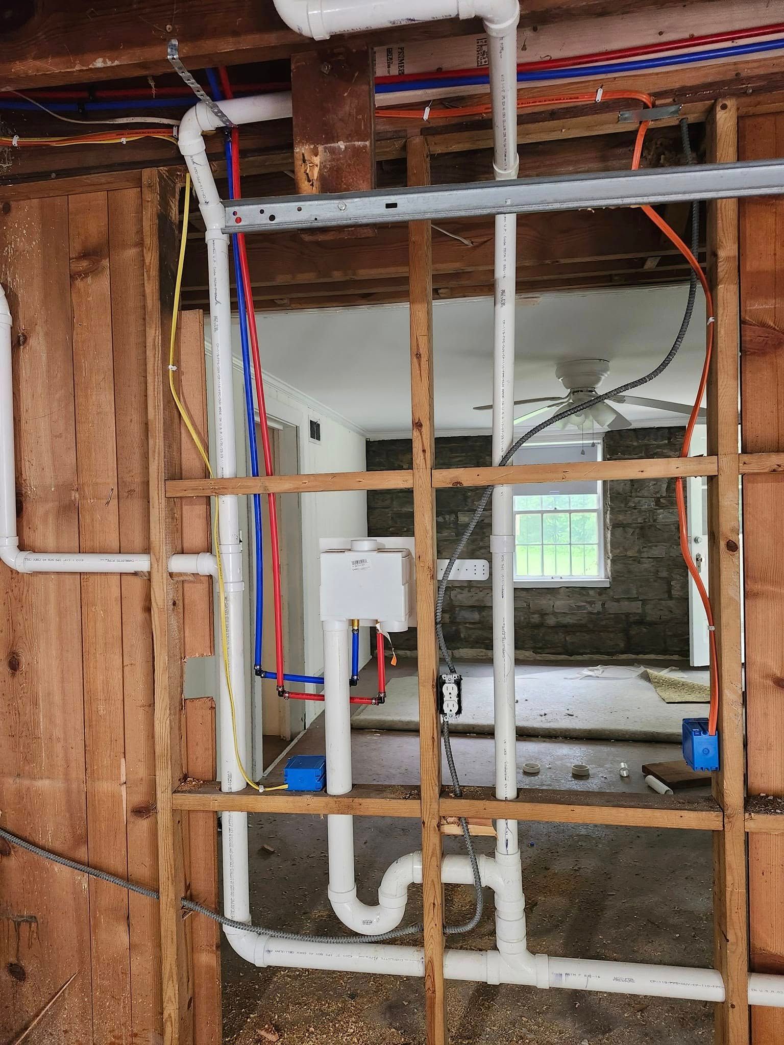 Plumbing and electrical work in a wall opening. White PVC pipes, blue and red tubes, and electrical wiring are visible.