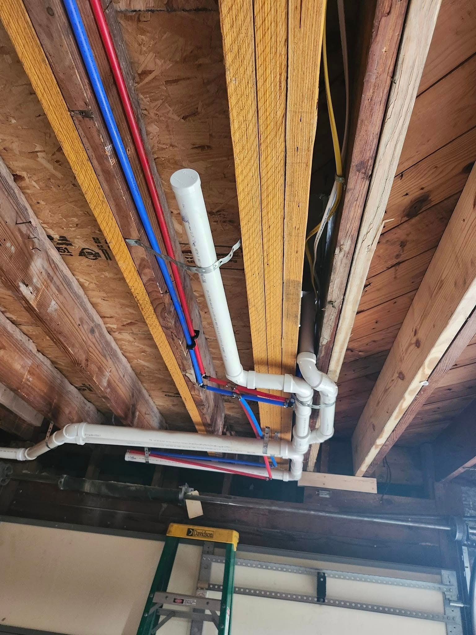 Plumbing pipes of various colors and a white vent pipe are installed under wooden beams in a ceiling.