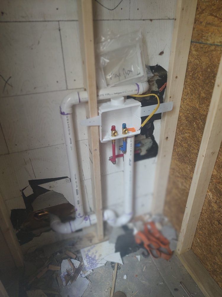 Plumbing for a washing machine in a wall. White PVC pipes and a white outlet box are visible.