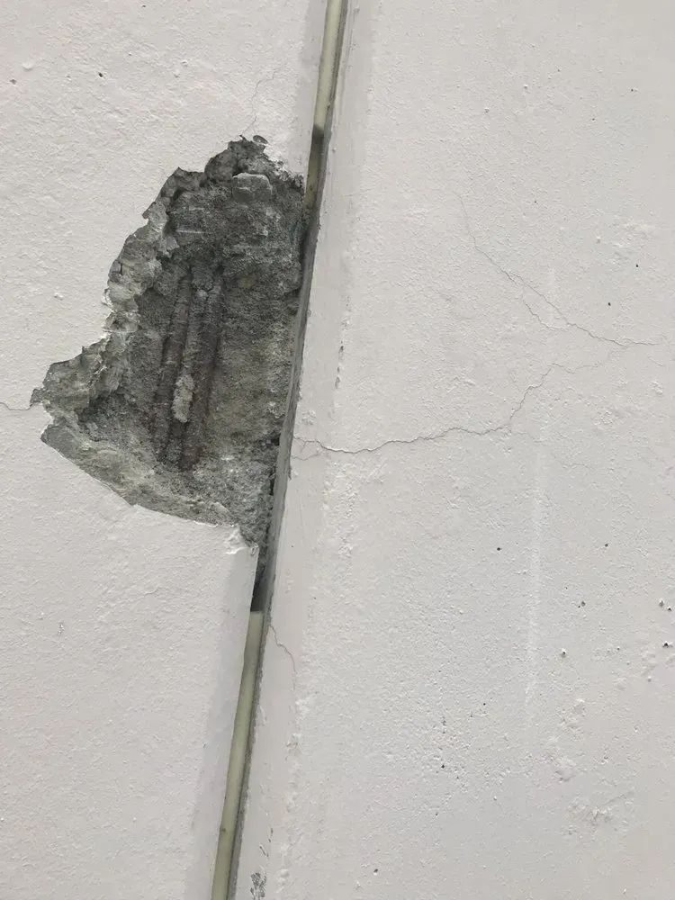 Damaged white wall reveals exposed concrete and metal reinforcement next to a vertical joint.