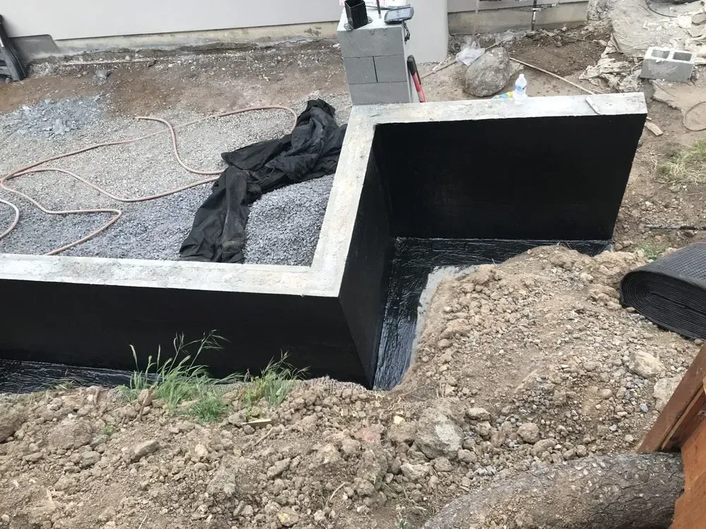 Concrete retaining wall covered in black waterproofing material, outdoors in a yard.