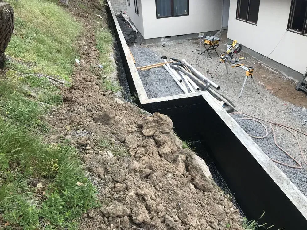 Excavated foundation wall next to a hillside. Black waterproof membrane visible. Construction site, light-colored buildings.