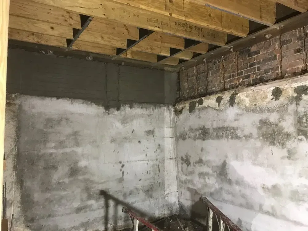 Basement interior with concrete walls, wooden beams overhead, and a staircase.