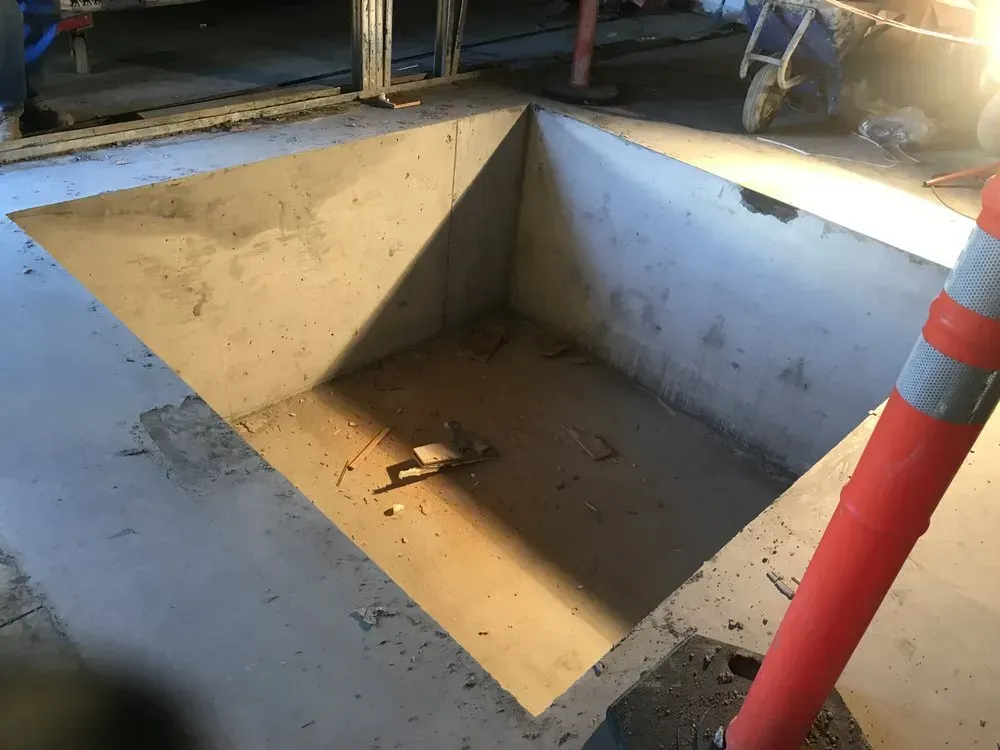 A concrete rectangular pit in a concrete floor, possibly for automotive work, with a traffic cone in the foreground.