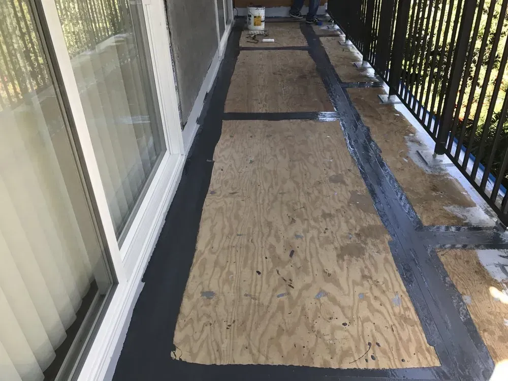 A narrow outdoor balcony under renovation, with gray sealant and plywood boards.