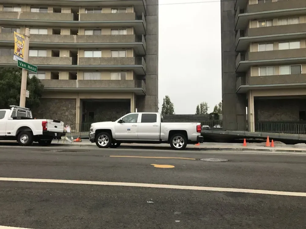 Two white pickup trucks parked near apartment buildings. A street separates them. Cloudy sky.