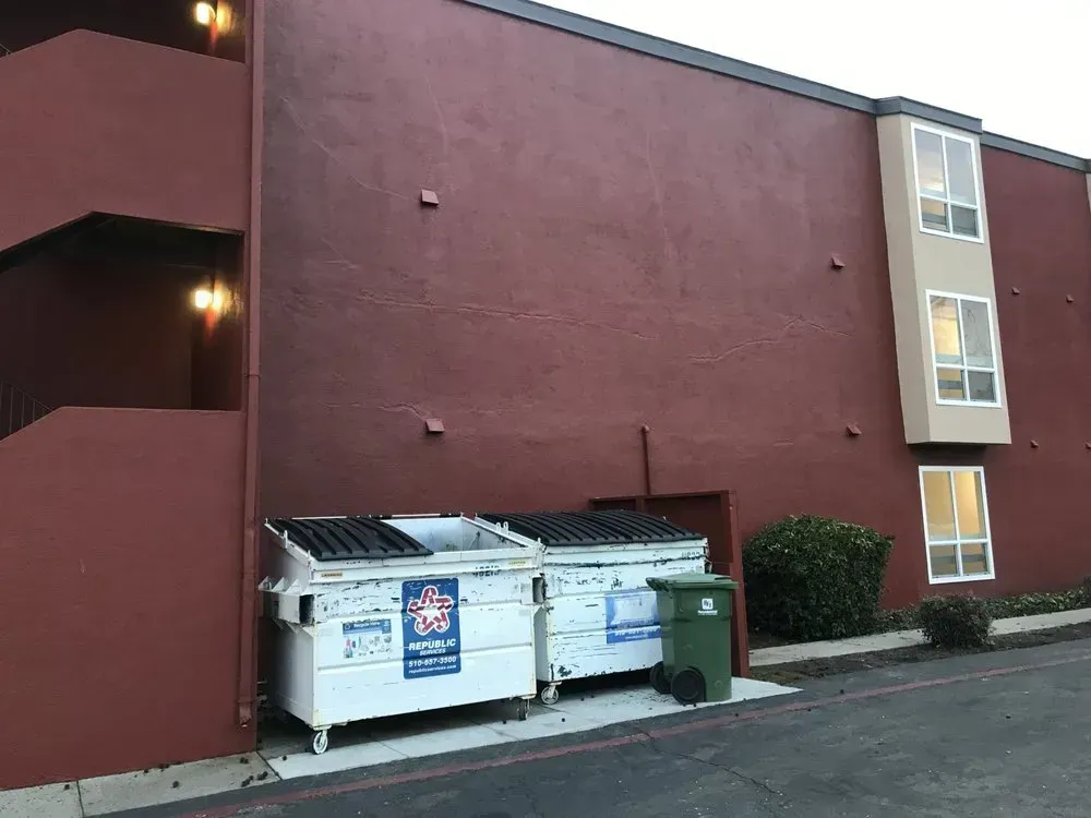 Two dumpsters next to a building with burgundy walls and a stairway. A green trash can is beside them.