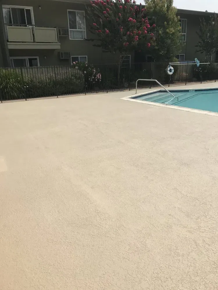 Poolside with tan concrete deck, fence, building, and clear blue water.