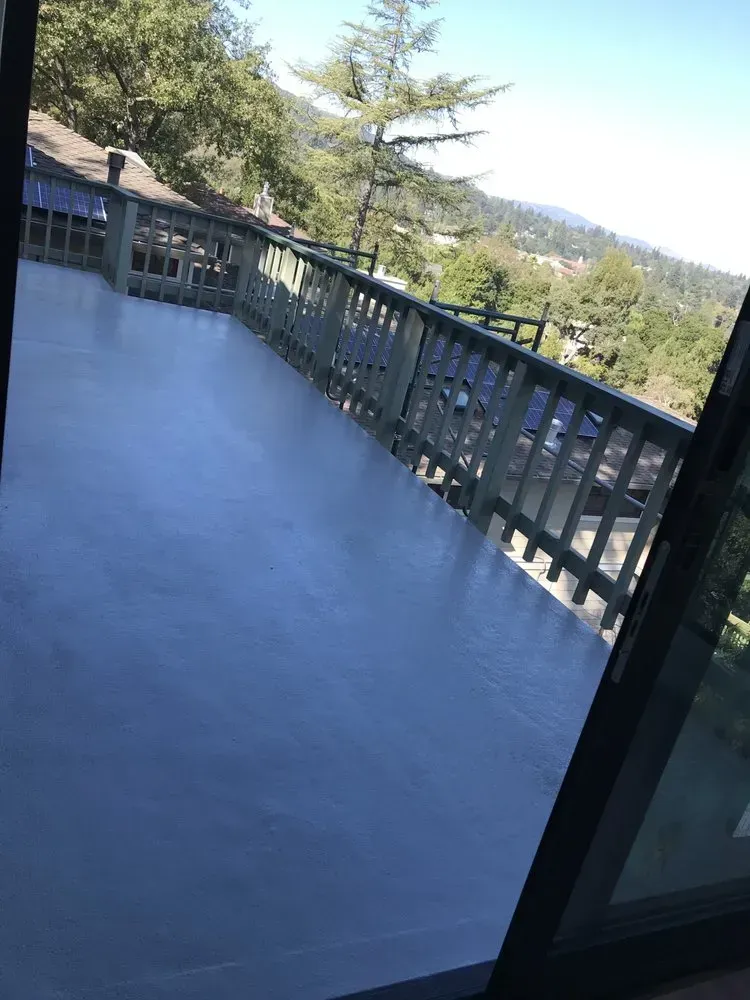 A gray deck with a railing overlooks a green valley and mountains, viewed from a glass door.