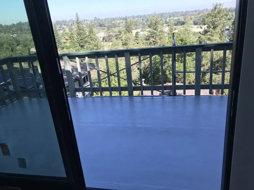 View of a blue-painted deck and railing overlooking trees and distant houses through a glass sliding door.