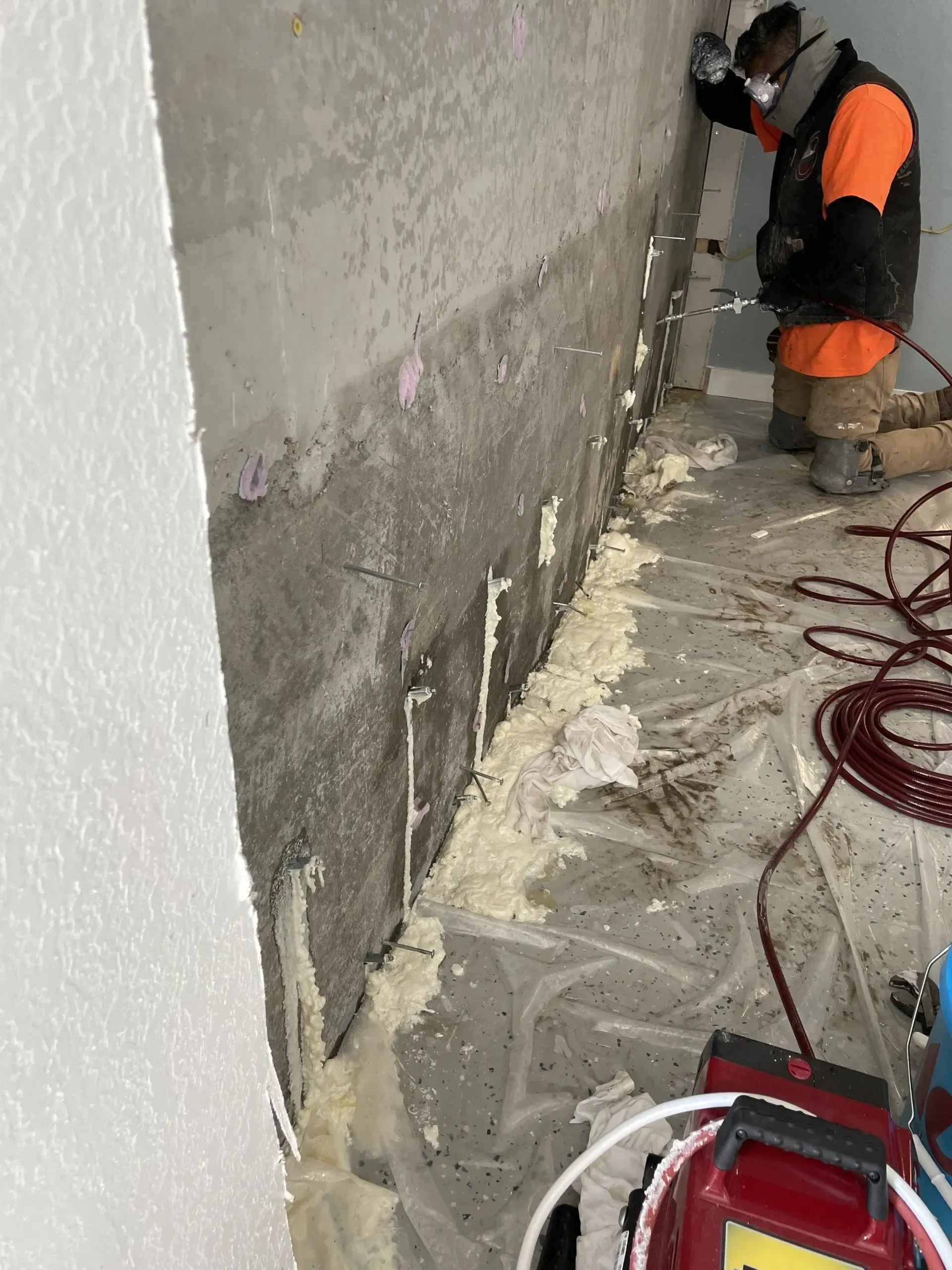 Construction worker spraying foam on a wall, with red wire and equipment on the floor.