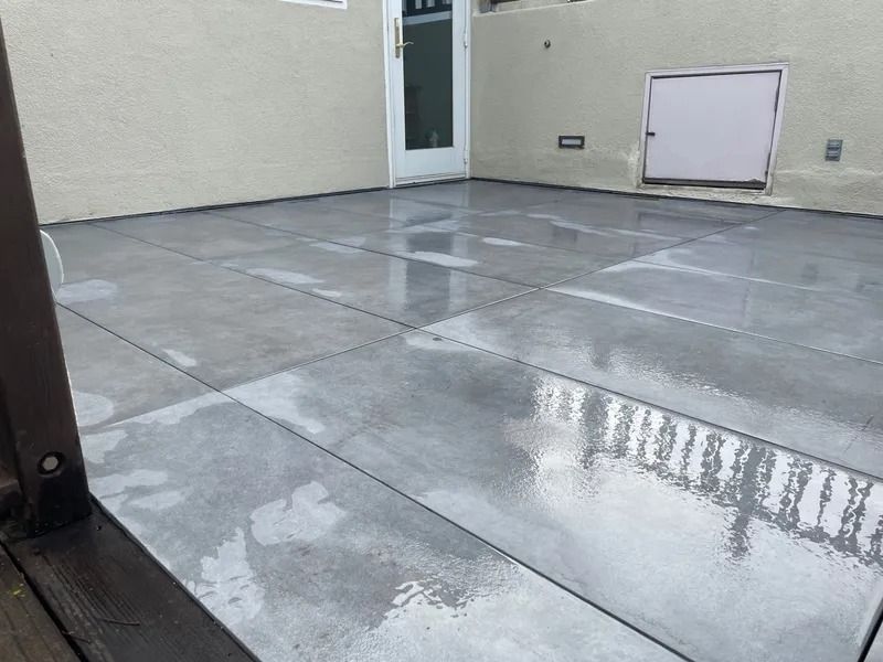 Gray patio with wet, rectangular tiles, near a wall with a door and a small white box.