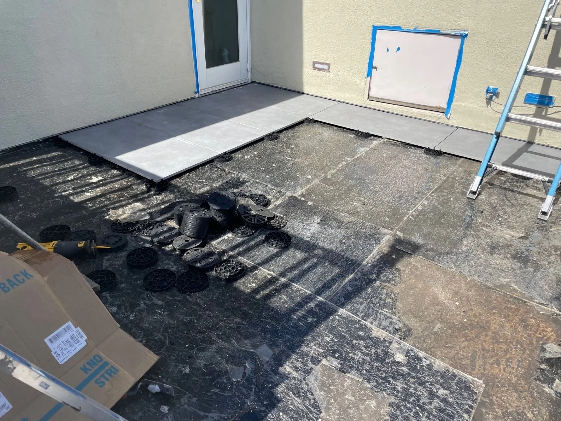 Patio under construction; gray tiles partially laid, black pedestal supports, ladder, cardboard box.
