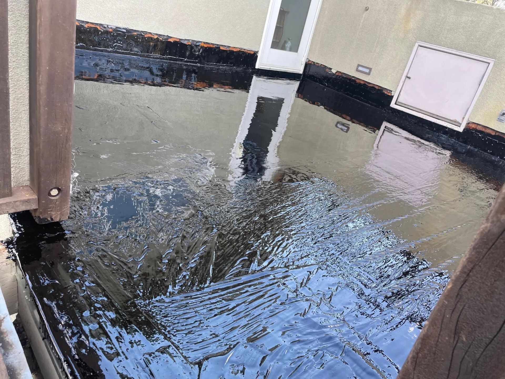 Black tar roof with standing water, reflecting a white door and beige wall.