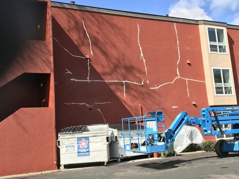 Red building exterior with multiple cracks; a dumpster and a blue lift are present.