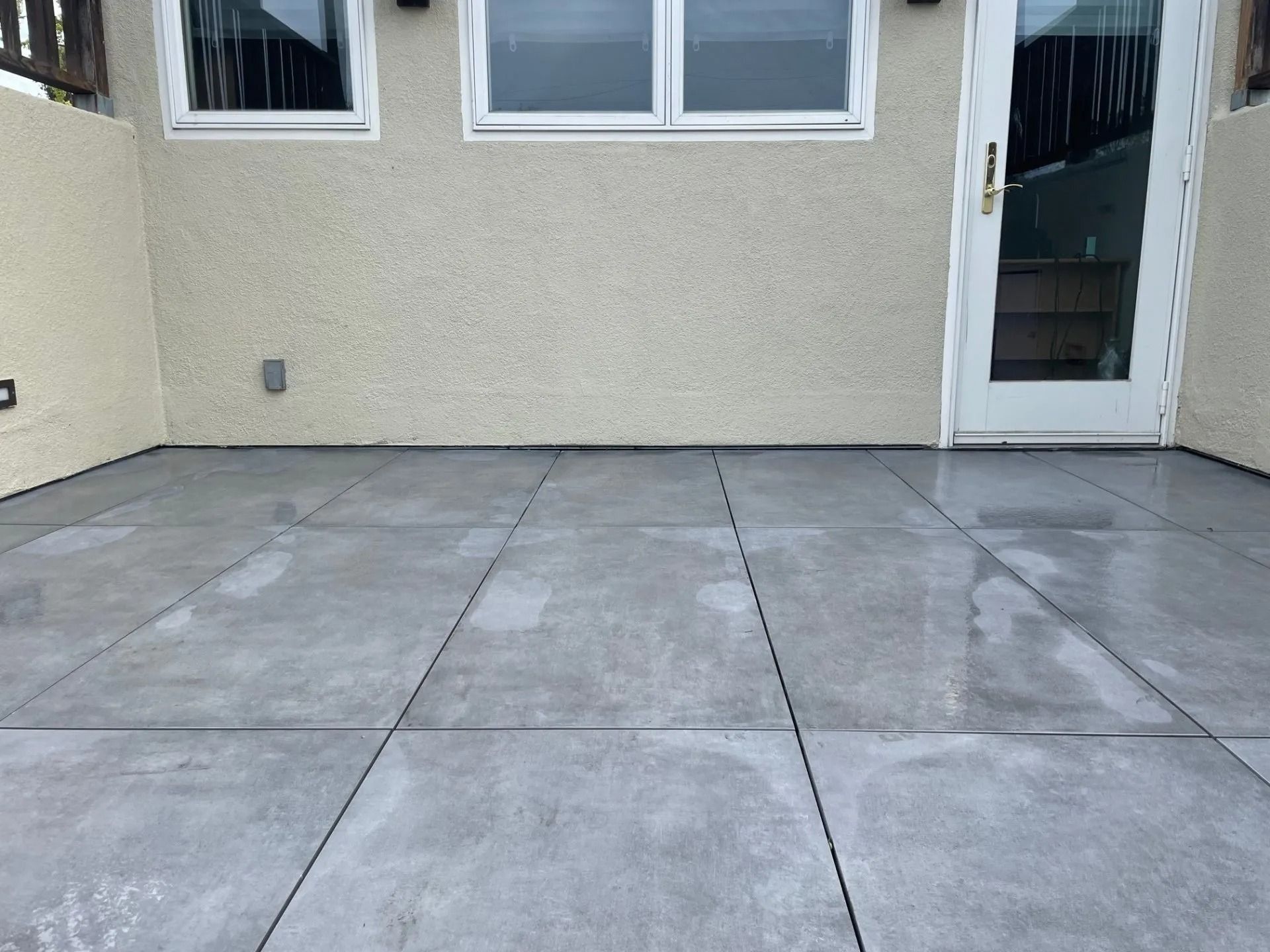 Gray concrete patio with wet patches, against a beige stucco wall with windows and a door.