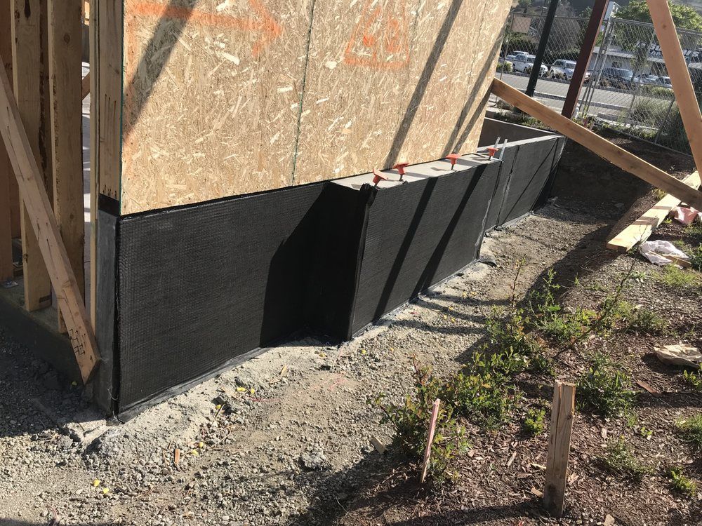 Black waterproofing on a building foundation, partially covered by soil and wood framing.