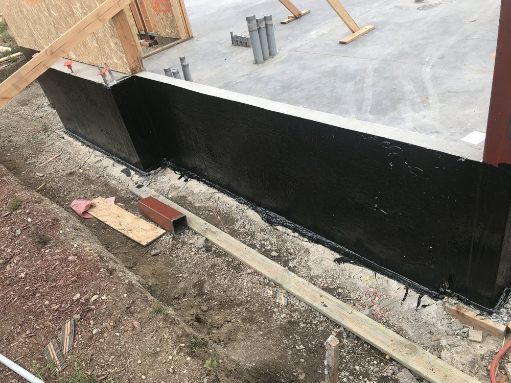 Black waterproof coating on a concrete foundation wall during construction. Soil, wood, and pipes are visible.