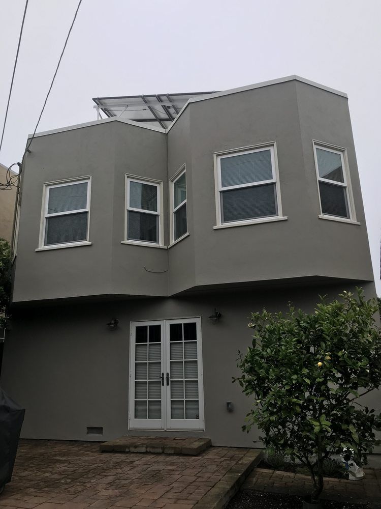 Two-story gray house with white-framed windows and double doors. Solar panels on the roof. Cloudy day.