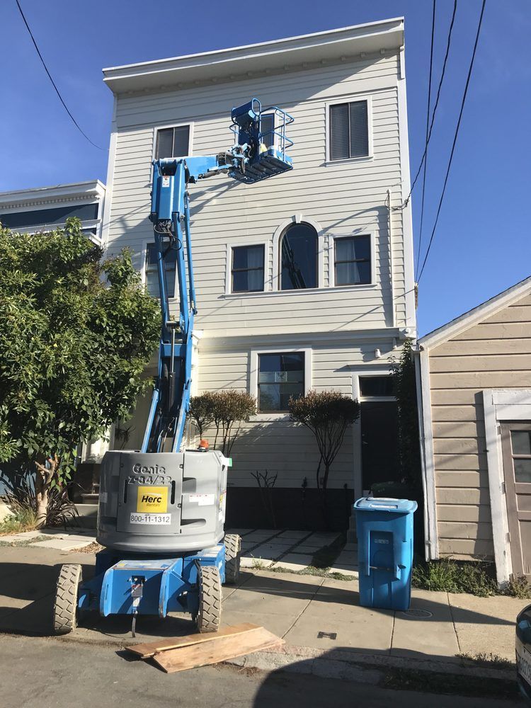 A blue lift servicing a three-story white house with dark-framed windows and a blue trash bin.