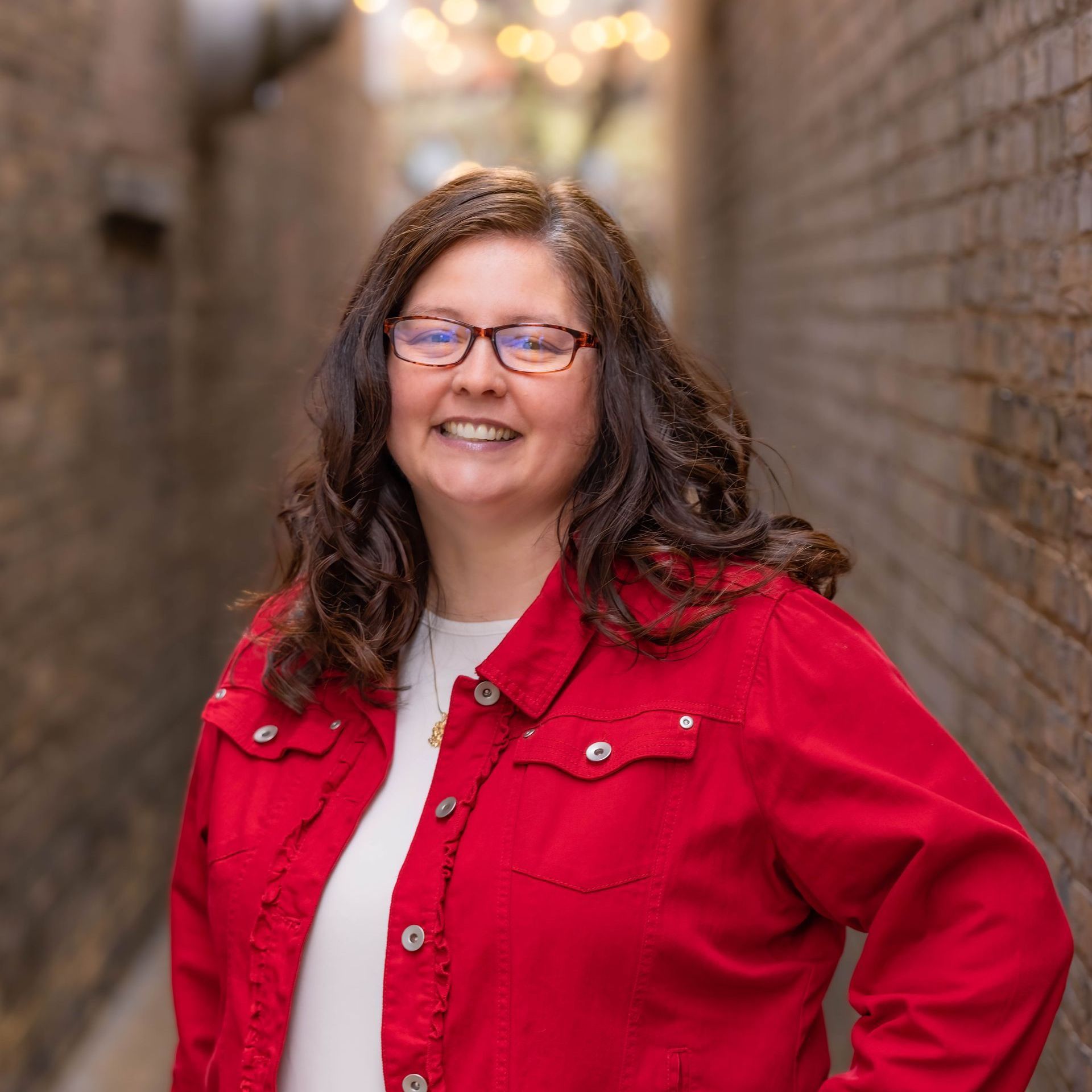 A woman wearing glasses and a red jacket is standing in front of a brick wall.