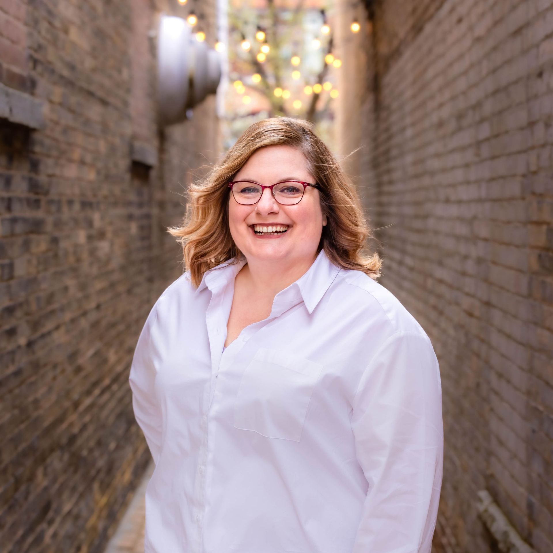 A woman wearing glasses and a white shirt is standing in a narrow alleyway.