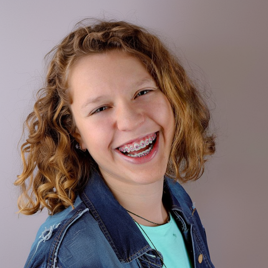 Smiling person with braces in a denim jacket and teal shirt, short curly brown hair against a gray background.