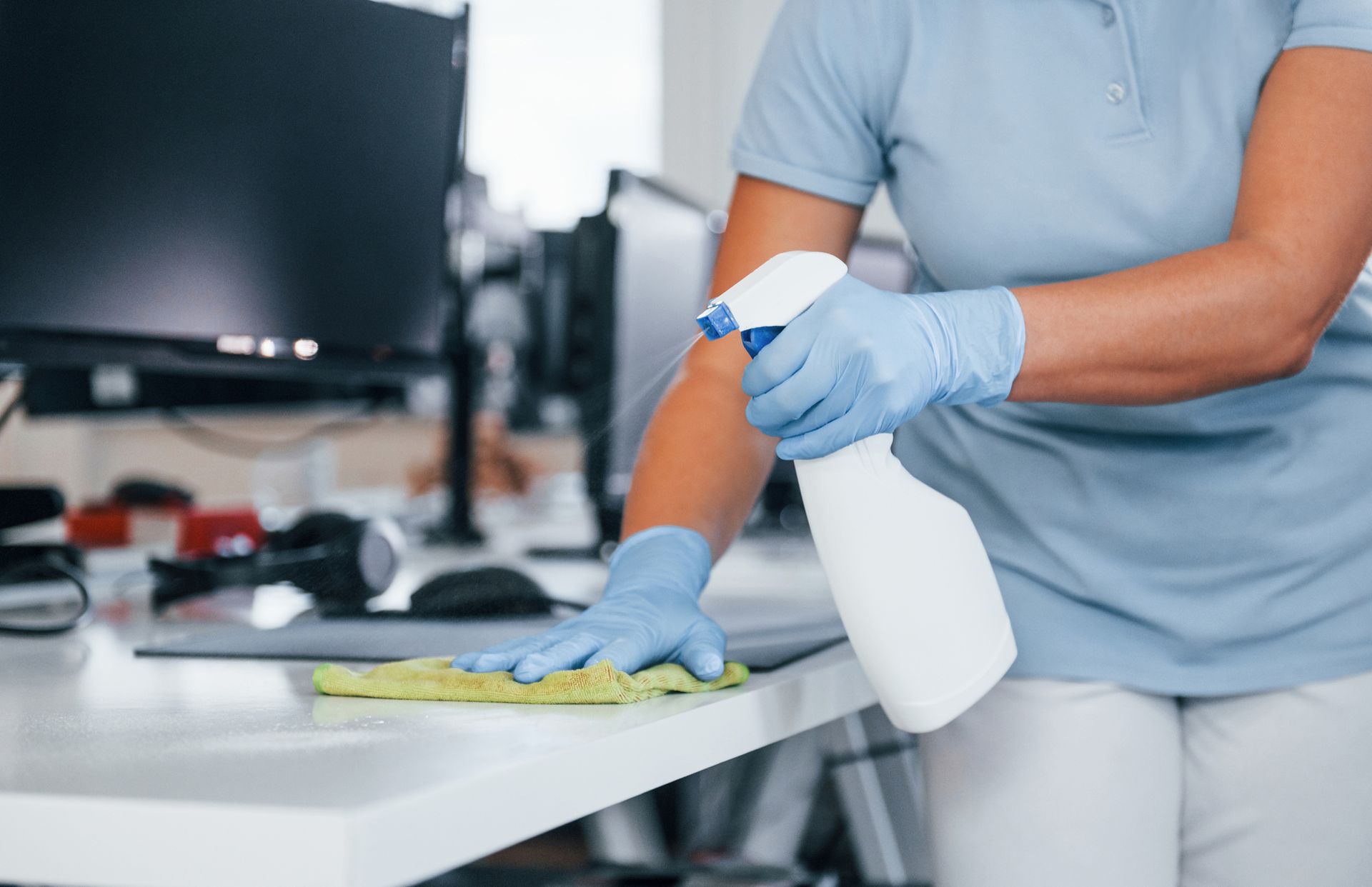 A woman is cleaning a desk with a spray bottle and a cloth.