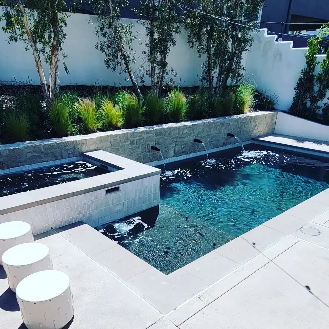 Modern outdoor pool with a spa. White concrete patio and wall with fountain jets. Green plants behind.