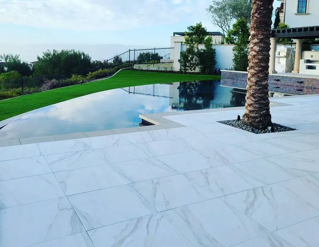 Pool with reflective water, green grass, stone patio, palm tree, and ocean view in background.