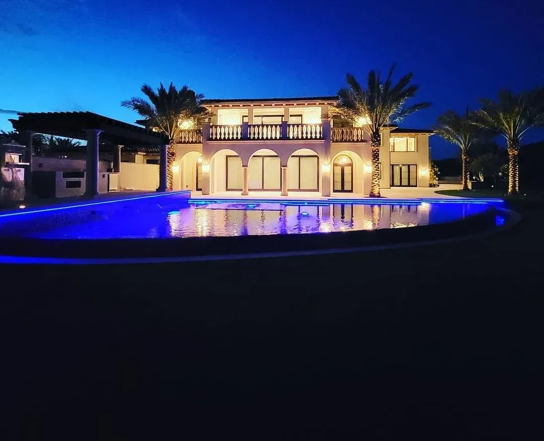 Night view of a mansion with a pool lit with blue lights. Palm trees and pergola.