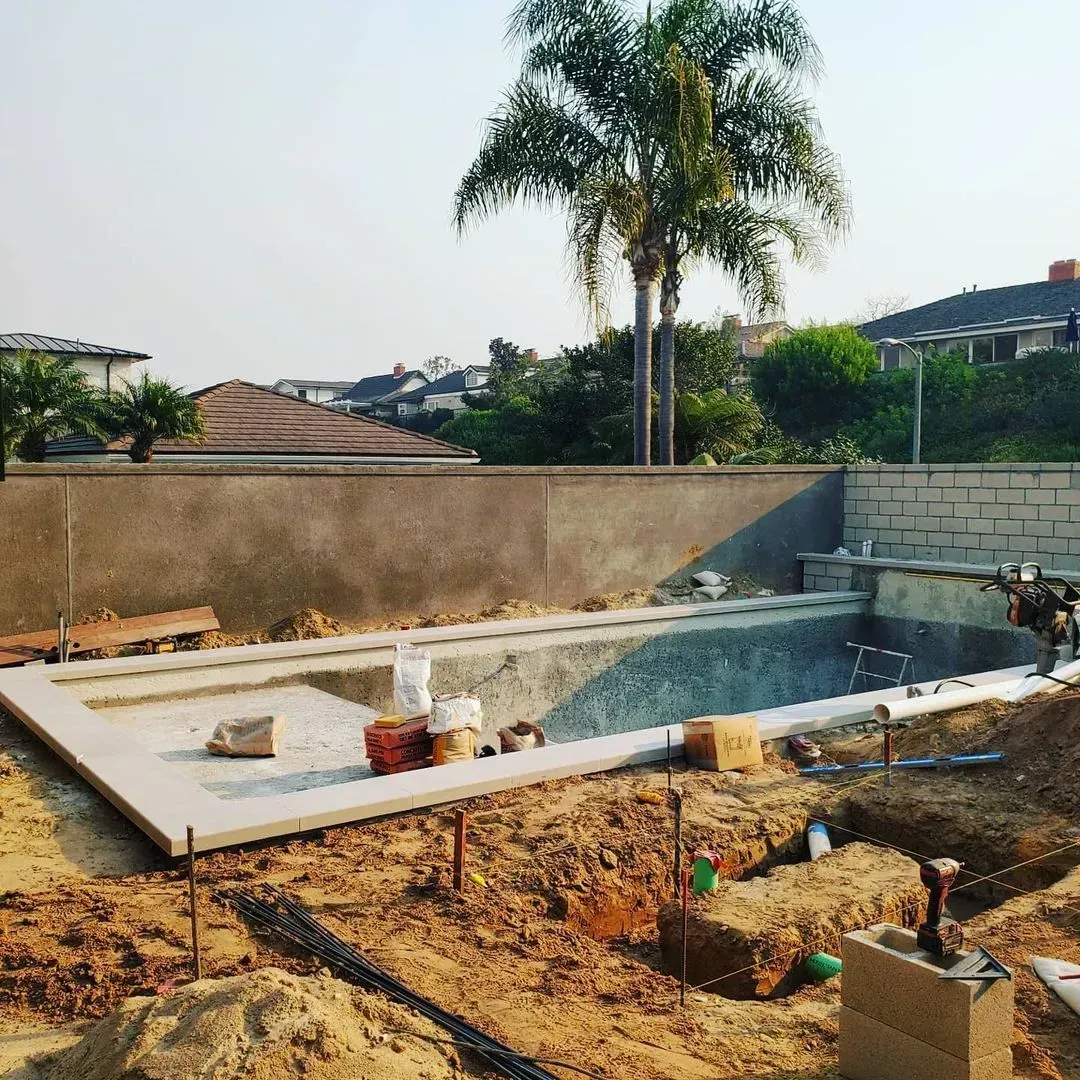 Pool construction site with concrete basin, dirt ground, and a retaining wall in a suburban backyard.